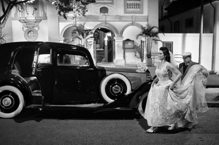 Fotografia poética e fotojornalistica de um casamento na capela da PUC a noiva em direção ao carro antigo e luxuoso com o motorista segurando a cauda de seu vestido branco enquanto o buque descansa no capo do carro