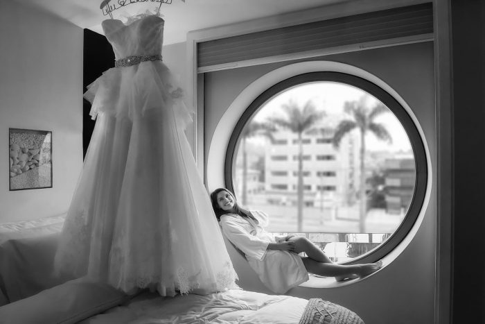 foto artística da noiva sentada na janela de seu quarto no hotel unique olhando seu grandioso vestido pendurado
